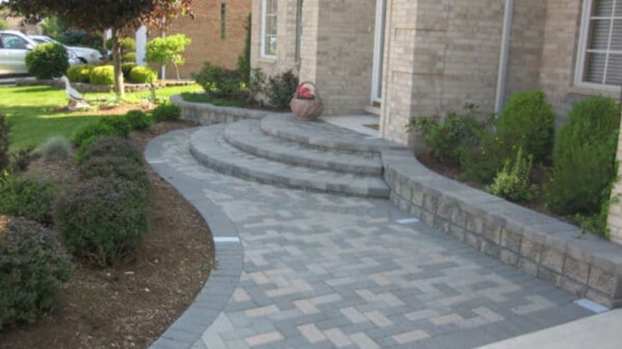 Curved stone walkway path with steps leading to a brick house entrance, bordered by garden beds with shrubs and plants.