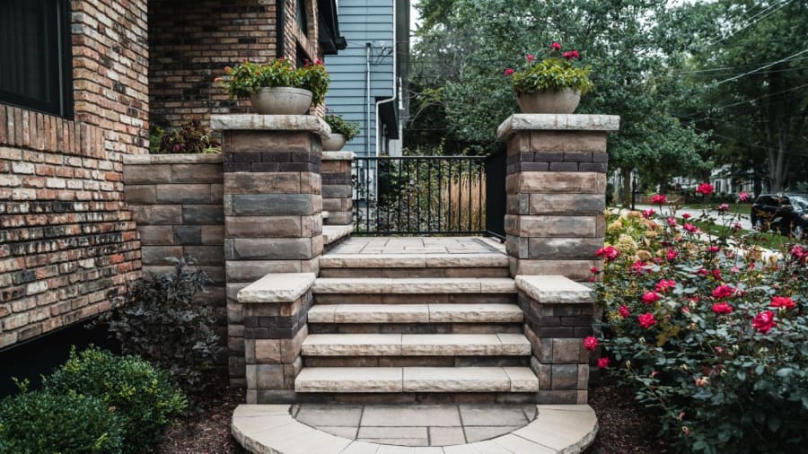 Four beige brick steps with stone pillars topped with flower pots on each side, red roses and greenery around the stairs, iron gate at the top.