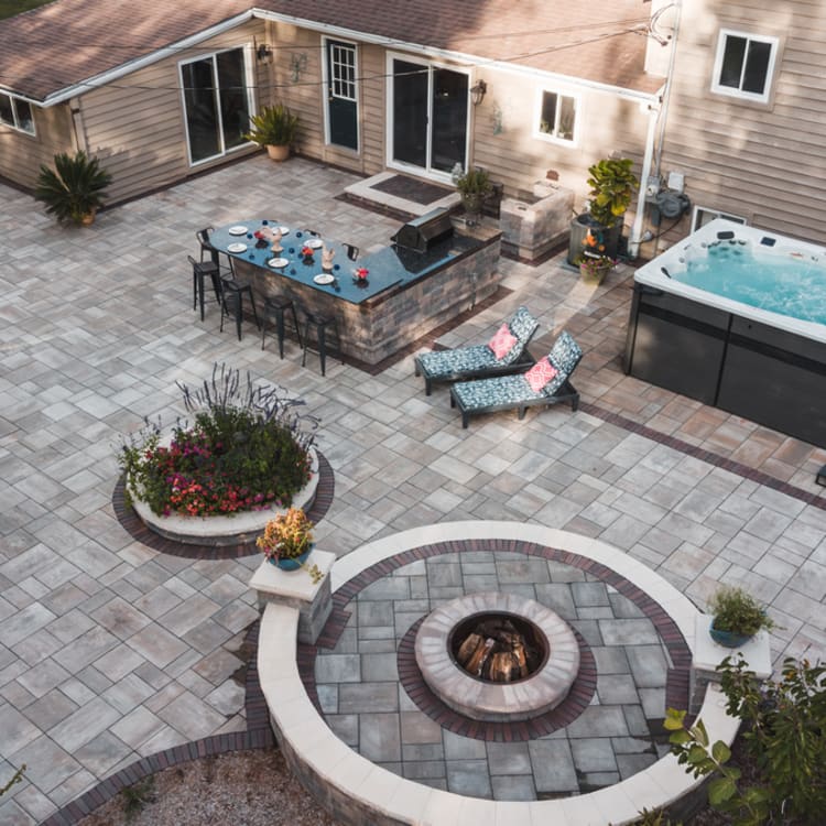 High-angle view of a large custom paver patio featuring an L-shaped outdoor kitchen, a circular fire pit with a masonry seat wall, and a large swim spa.
