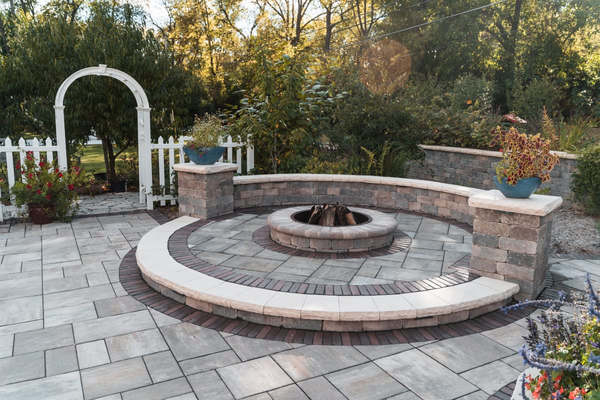 A circular stone fire pit on a paver patio with a curved seating wall, bordered by landscaping and a white garden arbor.